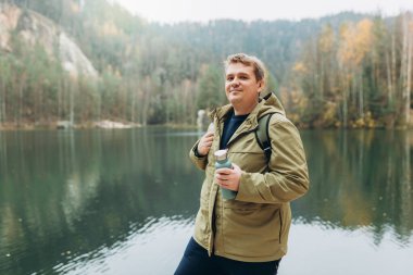 Sırt çantalı gülümseyen bir erkek gezgin görkemli gölün güzel manzarasının tadını çıkarıyor. Seyahat, macera. Etkin bir yaşam biçimi kavramı.
