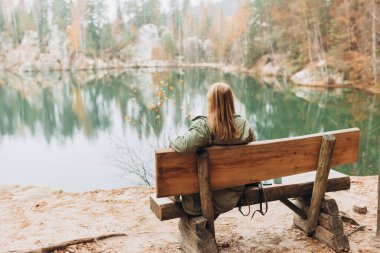30 'lu yaşlardaki yalnız sarışın kadın bir orman gölünün bankında oturuyor. Sonbahar manzarası, sakinlik, sükunet, seyahat konsepti. Gölün kıyısındaki bir kadın doğanın tadını çıkarıyor. Yüksek kalite fotoğraf.