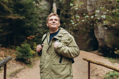 Sırt çantalı bir turist bir dağ ve ormanın arka planında duruyor. 30 'lu yaşlarda genç sarışın bir adam doğa yürüyüşü yapıyor. Seyahat, macera. Etkin bir yaşam biçimi kavramı.