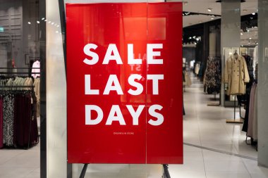 Giyim mağazasının önündeki indirim tabelası. Son günlerde indirimli. Vitrinde parlak kırmızı bir pankart var. Mağaza girişinde indirim bilgileri, alışveriş sezonu tanıtımı.