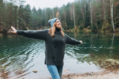 Gölün kıyısında elini kaldıran mutlu kadın. Genç otuzlarında kadın yürüyüşçü mutlu bir şekilde gülümsüyor, ormanda yürüyüş yapıyor, ılık bir sonbahar günü..