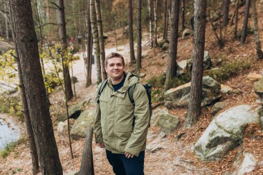 Sırt çantalı bir turist bir dağ ve ormanın arka planında duruyor. 30 'lu yaşlarda genç sarışın bir adam doğa yürüyüşü yapıyor. Seyahat, macera. Etkin bir yaşam biçimi kavramı.