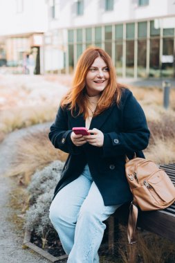 Mutlu kadın bankta oturuyor ve dışarıda telefonla konuşuyor. Kentsel, halk konsepti. Gülümseyen 30 'lu yaşlardaki kızıl saçlı kadın şehrin güneşli bir gününde, bahar zamanı akıllı telefondan mesaj atıyor..