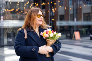 İlkbahar laleli güzel bir kadın çiçek buketi ile şehir caddesinde yürüyor. Dışarıda poz veren mutlu kız. Kentsel arka planda güzel bir kadının bahar portresi