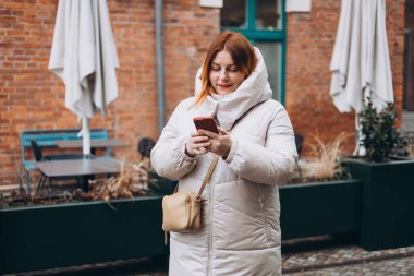 30 'lu yaşlardaki genç bir kadın akıllı telefondan şehirde yürürken mesaj yazıyor. Şehir yaşam tarzı konsepti. Sonbahar geldi. Etkileyici bir kadın şık kıyafetler giyip internette sohbet ediyor. Yüksek kalite fotoğraf.
