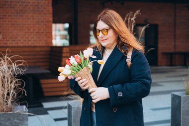 İlkbahar laleli güzel bir kadın çiçek buketi ile şehir caddesinde yürüyor. Dışarıda poz veren mutlu kız. Kentsel arka planda güzel bir kadının bahar portresi