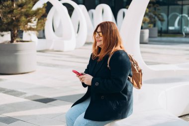 Mutlu kadın telefonda konuşuyor ve dışarıda dinleniyor. Kentsel, halk konsepti. Gülümseyen kadın, güneşli bir günde, ilkbahar vakti akıllı telefondan mesaj atıyor.