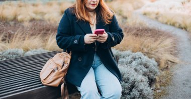 Mutlu kadın bankta oturuyor ve dışarıda telefonla konuşuyor. Kentsel, halk konsepti. Gülümseyen 30 'lu yaşlardaki kızıl saçlı kadın şehrin güneşli bir gününde, bahar zamanı akıllı telefondan mesaj atıyor..