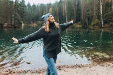 Gölün kıyısında elini kaldıran mutlu kadın. Genç otuzlarında kadın yürüyüşçü mutlu bir şekilde gülümsüyor, ormanda yürüyüş yapıyor, ılık bir sonbahar günü..