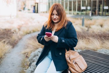 Mutlu kadın bankta oturuyor ve dışarıda telefonla konuşuyor. Kentsel, halk konsepti. Gülümseyen 30 'lu yaşlardaki kızıl saçlı kadın şehrin güneşli bir gününde, bahar zamanı akıllı telefondan mesaj atıyor..