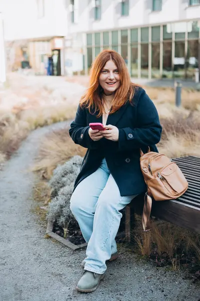 Mutlu kadın bankta oturuyor ve dışarıda telefonla konuşuyor. Kentsel, halk konsepti. Gülümseyen 30 'lu yaşlardaki kızıl saçlı kadın şehrin güneşli bir gününde, bahar zamanı akıllı telefondan mesaj atıyor..