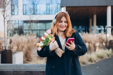 30 'lu yılların beyaz bayan öğrencisi kablosuz kulaklıklı, telefon dinlemeli, müzik dinlemeli, üniversite kampüsünde ses dersinin keyfini çıkarmalı, açık havada..