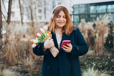 30 'lu yılların beyaz bayan öğrencisi kablosuz kulaklıklı, telefon dinlemeli, müzik dinlemeli, üniversite kampüsünde ses dersinin keyfini çıkarmalı, açık havada..