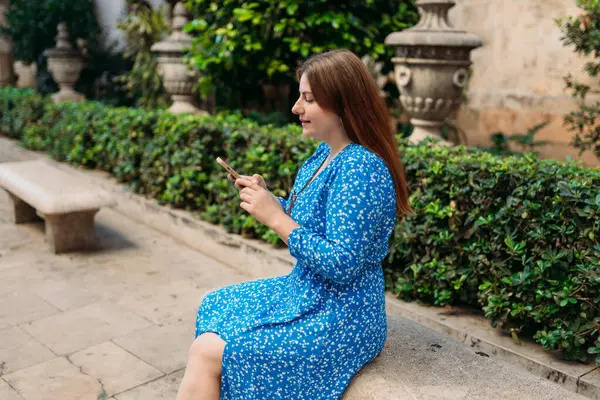 Eski bankta oturan ve dışarıda telefonla konuşan mutlu kadın. Güneşli bir günde şehrin sokaklarında gülümseyen kızıl saçlı bir kadın akıllı telefondan mesaj atıyor.