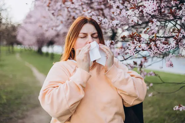 Parkta çiçek açan ağaçların arasında burun sileceği olan genç kızıl saçlı bir kadın. Hasta kadınların portresi beyaz dokuya hapşırır, burun kıvrımlarından muzdariptir. Soğuk algınlığı veya alerji belirtileri