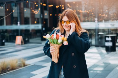 Mutlu 30 'lu yaşlardaki laleli, çiçek buketli, telefonda konuşan ve dışarıda mutlu görünen kadın. Şehir yaşam tarzı konsepti. Bahar geldi. Modern şehir caddesinde yürüyen güzel bir kadın..