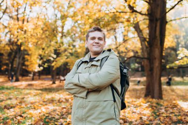 Autumn Park 'ta rahatlayan mutlu 30' lu yaşlarda bir adam. Gezgin. Düşme zamanı. Dışarıda dikilen, günlük kıyafetli, gülümseyen adam.