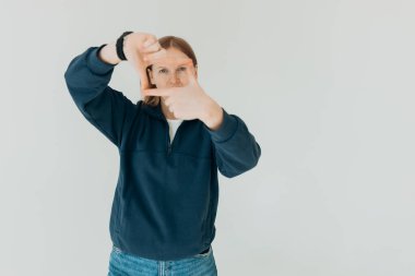 Genç ve güzel bir kadın izole edilmiş beyaz arka planda duruyor gülümsüyor ve elleri ve parmakları gülen yüzlü bir çerçeve yapıyor. Yaratıcılık ve fotoğrafçılık kavramı