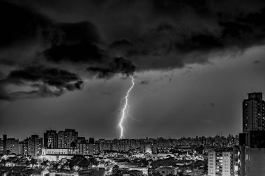 Şehir manzarası üzerinde bir yıldırım fırtınasının dramatik siyah beyaz fotoğrafı. Sürgüler karanlık bulutları ve binaları aydınlatır ve yoğun bir atmosfer yaratır. Şehir ışıkları, doğanın gücünü gösteren uğursuz gökyüzü ile tezat oluşturuyor..