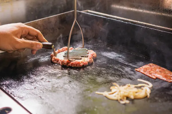 Adam ızgarada spatula ile biftek köftesi eziyor.