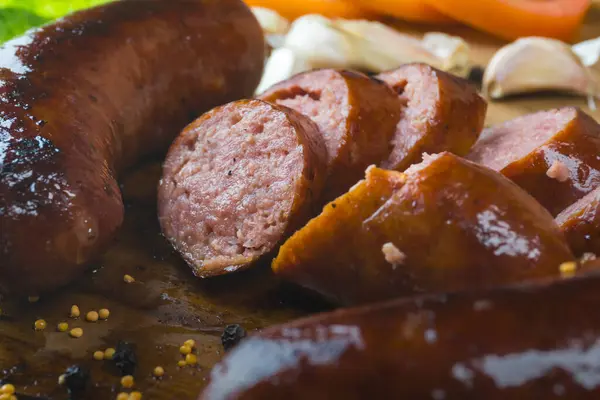 Stock image Grilled juicy sausages that have been sliced after cooking, on a wooden cutting board. Lettuce, tomato and garlic in the background