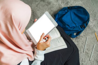Müslüman bir kadın oturma odasında tablet kullanarak ders çalışıyor.