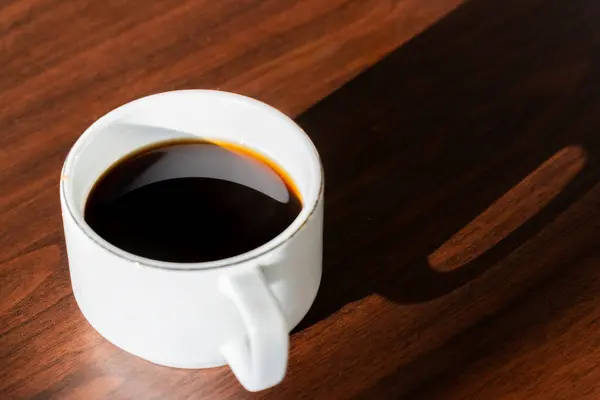 Sabah kahvesi beyaz fincanda ve fincandan uzun bir gölge tahta masada.