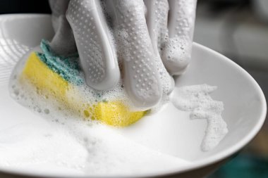 Lastik eldiven giyen eller sünger kullanarak bulaşık yıkıyor.