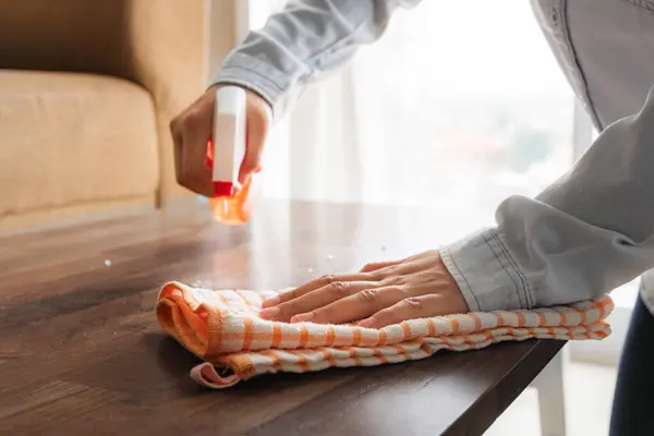 Bir kadın oturma odasındaki ahşap masanın yüzeyini temiz bir bez, sprey bir şişe ve ev temizleme konseptiyle temizliyor.