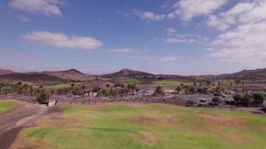 Fuerteventura 'da içi palmiye ağaçları ve volkanik dağlarla kaplı bir yol olan manzaralı bir golf sahasının hava görüntüleri. Gün ışığında yakalanan sakin sinema hareketleriyle adaların yemyeşil ve kurak arazilerinin eşsiz bir karışımını sergiliyor.. 
