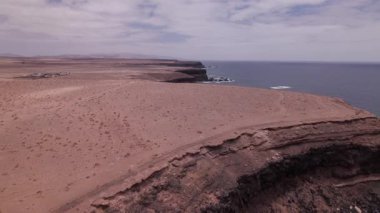 Fuerteventura 'daki Playa de la Escalera' nın sert, rüzgarlı kıyılarında uçan bir hava videosu. Kırmızı volkanik arazi, kayalık yamaçlar ve çarpan dalgalar ham ve güzel bir ada manzarası oluşturur..