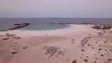 İnsansız hava aracı görüntüleri Fuerteventura 'daki La Concha Sahili' ni turkuaz su, beyaz kum ve siyah volkanik kayalarla gösteriyor. Gün ışığında adanın eşsiz kıyı şeridini ve doğal kontrastı vurguluyor..