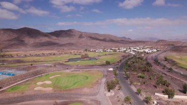 Hava aracı görüntüleri Fuerteventura 'nın engebeli volkanik manzarasına karşı yemyeşil bir golf sahası ve palmiye çizgili bir yol olduğunu gösteriyor. Sahne yeşil çim yolları, kurak çevreler ve açık mavi gökyüzünün altındaki uzak dağlar arasındaki zıtlığı yakalar..