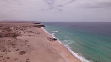 Fuerteventura 'daki Playa del Aljibe de la Cueva üzerinde geniş açılı drone görüntüleri. Volkanik kayalıklar ve açık gökyüzü tarafından çerçevelenmiş geniş kumlu sahilde yalnız bir paraşütçü duruyor..