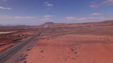 Hava aracı Fuerteventura, Kanarya Adaları 'nın kırmızı çöl arazisinden geçen bir yolu takip ediyor. Manzarada volkanik toprak, minimum bitki örtüsü ve ufukta engebeli dağlar bulunur.. 