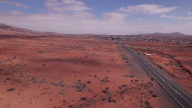 Fuerteventura 'nın kırmızı volkanik çölü boyunca uzanan uzak bir yolun hava görüntüleri. Kuru toprak ve asgari bitki örtüsüyle çevrili İHA, dağlar ile uçsuz bucaksız, boş araziyi yakalar..