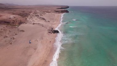 Hava aracı videosu Fuerteventura 'da Playa del Aljibe de la Cueva' yı gösteriyor. Deniz kenarına yakın bir yerde paraşütçü bir adam var. Engebeli kayalıklar ve turkuaz su çarpıcı bir manzara yaratır..