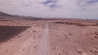 Tozlu bir yolun üzerinde uçan yüksek irtifa drone klipleri Fuerteventura 'daki El Cotillo kıyısındaki kırmızı volkanik toprağı kesiyor. Videoda adanın engebeli, kuru arazisi ve uzak deniz manzarası gösteriliyor..