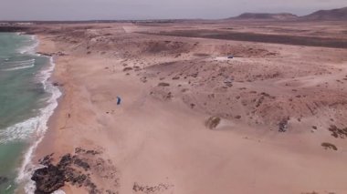 Fuerteventura 'daki Playa Mallorquines üzerindeki hava görüntülerinde sahile vuran dalgalar ve sahilde deniz paraşütü hazırlayan bir kişi görülüyor. Engebeli kayalıklar ve geniş kumlar manzarayı tamamlıyor.