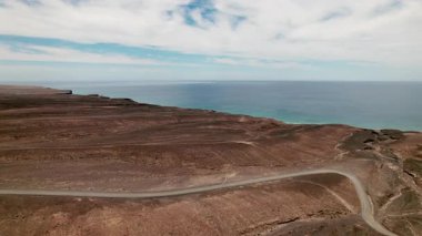 Kuru sıradağların üzerinden uçan bir dron görüntüsü ve Faro Punta de Jandia 'ya giden tozlu yol. Fuerteventura 'nın en güney bölgesinin ham ve doğal ortamını yakalar.. 