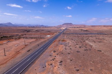 Fuerteventura 'daki dramatik kırmızı volkanik manzarayı kesen asfaltlı yolun geniş manzarası. Sahne, çorak güzelliği, derinliği ve seyahat potansiyelini yakalar..