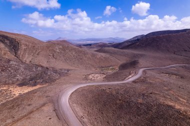 Fuerteventura, Kanarya Adaları 'ndaki kırmızı volkanik tepeleri ve çorak araziyi kesen kavisli toprak yolun hava manzarası. Mavi gökyüzünün altında vahşi, uzak bir manzara..