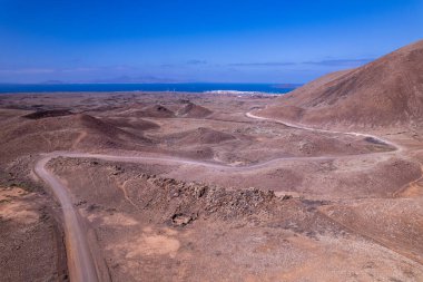 Fuerteventura 'daki engebeli volkanik tepelerin arasından geçen dolambaçlı toprak bir yolun yüksek açılı drone fotoğrafı. Kırmızı tonlar ve kuru jeoloji bu kurak araziyi tanımlar.