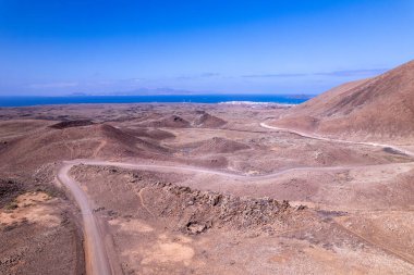 Fuerteventura 'nın kırmızı volkanik arazisindeki dolambaçlı toprak bir yolun drone görüntüsü. Çerçevenin hakimi engebeli manzara ve uzak kuru tepeler..