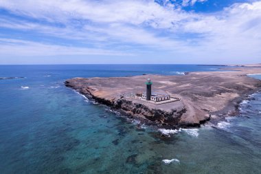Atlantik Okyanusu 'nun kristal berrak turkuaz sularının üzerindeki kayalık uçurumlarda gururla duran ikonik Faro de Jandia deniz fenerini gösteren muhteşem hava çekimi. Kırmızı volkanik arazi ve deniz arasındaki zıtlık çarpıcı bir manzara yaratır.. 