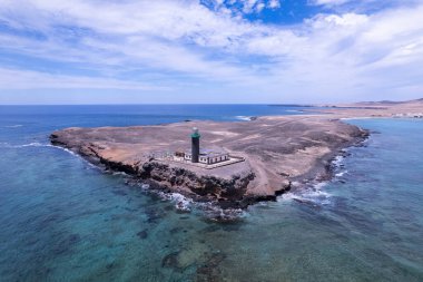 Geniş açılı çekimde, Atlantik Okyanusu 'nun turkuaz sularına bakan engebeli kayalık bir çıkıntıya tünemiş ikonik Faro de Jandia deniz fenerini yakalıyoruz. Görüntü volkanik arazinin ve açık mavi gökyüzünün katıksız güzelliğini gösteriyor..