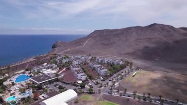 Fuerteventura 'daki Playitas Sahili yakınlarındaki büyüleyici sahil köyünü gösteren muhteşem hava görüntüleri. Görüntü modern mimari, siyah kum plajı ve turkuaz suların karışımını sergiliyor, seyahat fotoğrafçılığı için mükemmel.. 