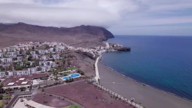 Fuerteventura 'daki Playitas Sahili yakınlarındaki büyüleyici sahil köyünü gösteren muhteşem hava görüntüleri. Görüntü modern mimari, siyah kum plajı ve turkuaz suların karışımını sergiliyor, seyahat için mükemmel, emlak fotoğrafçılığı. 