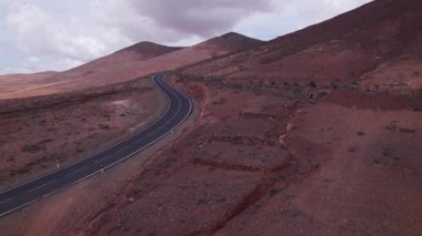 Fuerteventura 'daki dramatik kırmızı volkanik manzarayı kesen asfaltlı yolun geniş manzarası. Sahne, çorak güzelliği, derinliği ve seyahat potansiyelini yakalar..