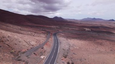 Fuerteventura 'daki dramatik kırmızı volkanik manzarayı kesen asfaltlı yolun geniş manzarası. Sahne, çorak güzelliği, derinliği ve seyahat potansiyelini yakalar..